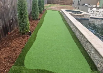 Backyard artificial turf strip beside pool with newly planted evergreens
