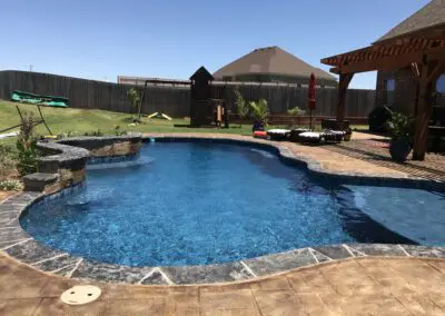 Backyard freeform blue pool with spa, stone coping, pergola and lounge chairs