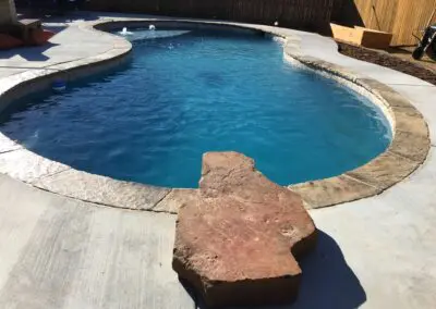 Concrete pool deck with curved blue swimming pool and large red stepping stone