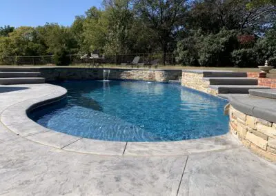 Curved backyard pool with stone coping, waterfall feature, and surrounding trees
