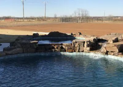 Backyard freeform pool with rock waterfall and raised spa, rural field background