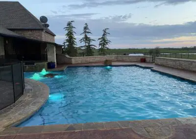 Backyard illuminated swimming pool with stone deck and twilight sky