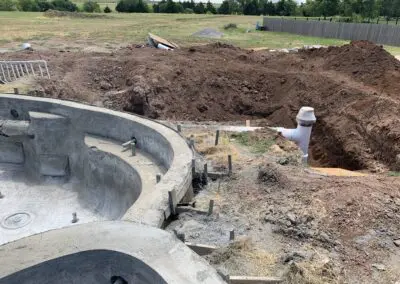 Concrete pool shell under construction beside large excavated backyard with plumbing