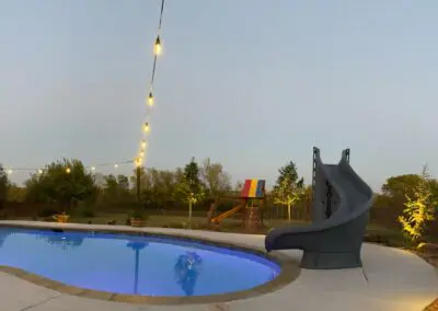 Backyard evening pool with spa, slide, string lights, and playset