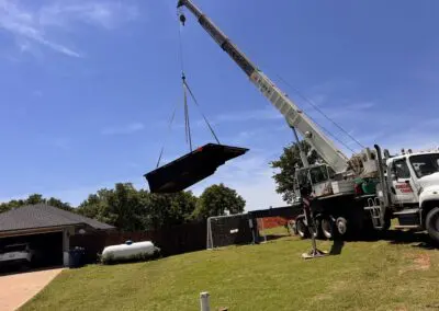 Crane lifting a black inground pool shell over a backyard toward house