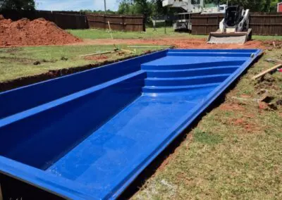 Blue fiberglass swimming pool shell being installed in backyard with crane and skid-steer