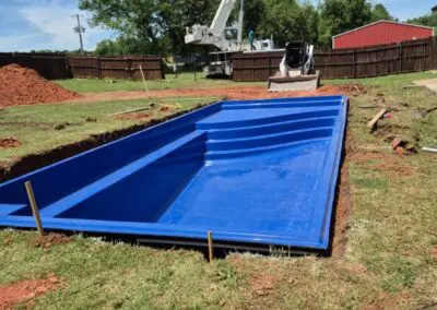 Blue fiberglass inground pool shell being installed in backyard with crane and skid-steer