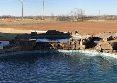 Backyard freeform pool with rock waterfall and raised spa, rural field background