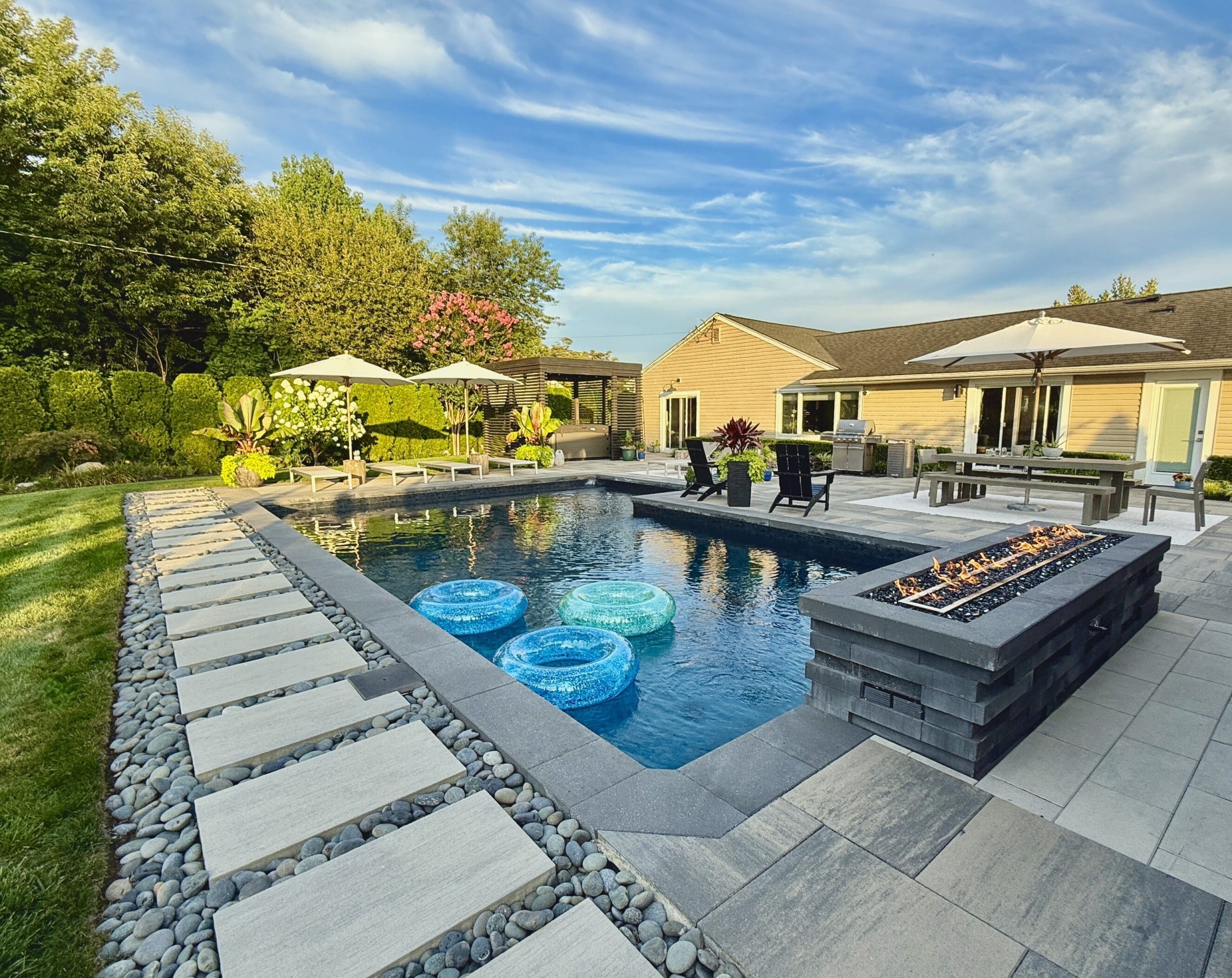 Modern backyard pool with pavers, fire pit, umbrellas, deck seating, and floats